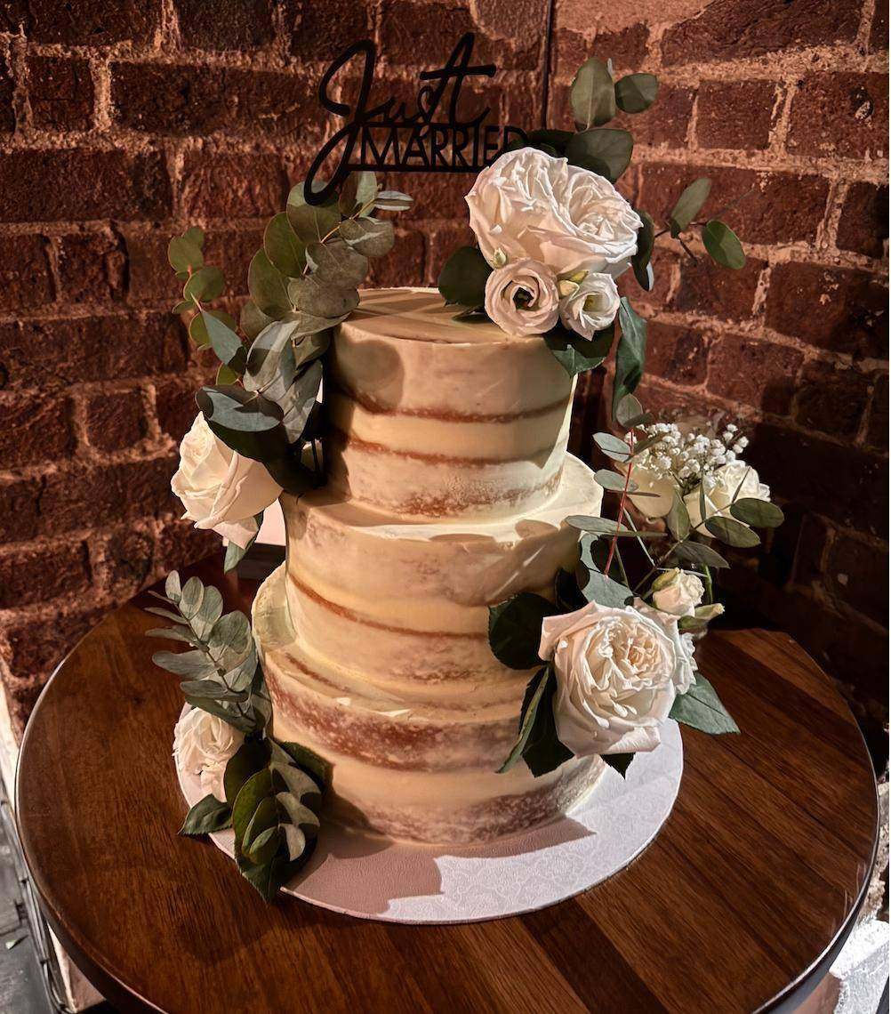 Semi-naked wedding cake decorated with white roses and a "Just Married" topper